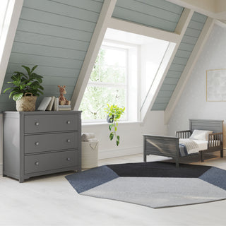 A cozy, modern kids’ room with slanted ceilings features the Child Craft Woodland Toddler Bed in Brushed Pebble with rails, a matching dresser, a potted plant, toys, books, a geometric rug, and a sunlit window.