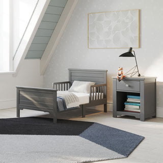A modern toddler bedroom features the Child Craft Woodland Toddler Bed in Brushed Pebble with white bedding, a gray nightstand and lamp, a stuffed animal, a book, patterned wallpaper, a large window, a geometric rug, and toddler bed rails for safety.