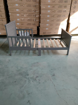 The Child Craft Woodland Toddler Bed in Brushed Pebble, designed for a crib mattress and featuring wooden slats, is shown partially assembled on a concrete floor before stacked boxes in a warehouse.