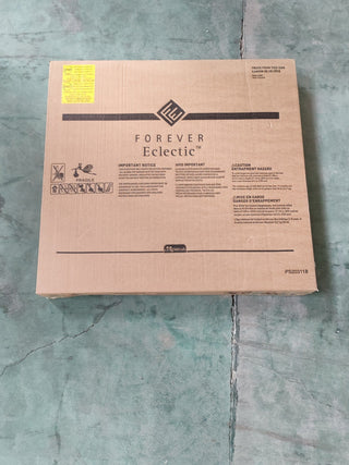 A sealed cardboard box labeled Child Craft rests on a greenish tiled floor. The box may contain a Woodland Toddler Bed, Brushed Pebble, and displays handling icons, warnings, and a yellow sticker in the top left corner.