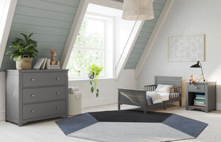 A cozy, light-filled kids room features the Child Craft Woodland Toddler Bed in Brushed Pebble with matching furniture, a round geometric rug, plants, a giraffe toy, and blue-gray walls with white trim beneath a sloped ceiling.