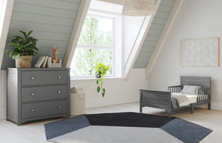 A cozy attic nursery with slanted ceilings features the Child Craft Woodland Toddler Bed in Brushed Pebble with secure rails, a matching dresser, potted plants, giraffe toy, books, a large window, and a geometric rug on light wood floors.