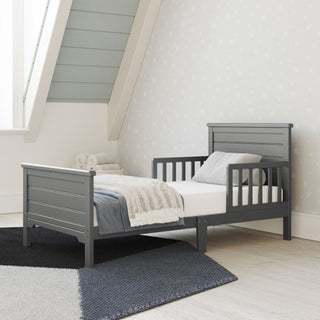 The Child Craft Woodland Toddler Bed in Brushed Pebble, featuring sturdy rails, pairs with a cozy crib mattress in a bright room with star-patterned wallpaper, a window, and folded towels at the head of the bed.
