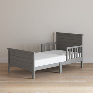A Child Craft Woodland Toddler Bed in Brushed Pebble with side rails, slatted headboard and footboard, and a white crib mattress is set on a light wood floor against a plain beige wall.