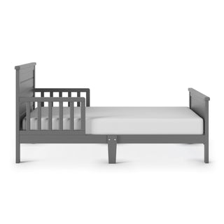 The Child Craft Woodland Toddler Bed in Brushed Pebble, shown from the side with toddler bed rails on one side and a white crib mattress, is displayed against a plain white background.