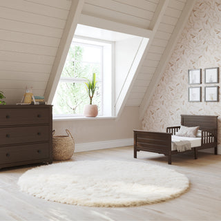 A cozy, light-filled nursery with a peaked ceiling features the Child Craft Woodland Toddler Bed in Brushed Truffle, a matching dresser, round white rug, woven basket, potted plant, and framed pictures on patterned wallpapered walls.