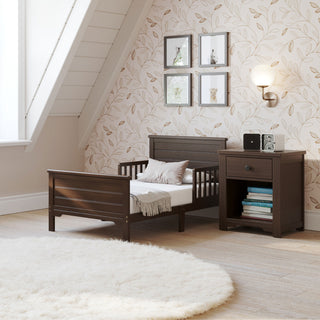 A cozy, modern toddler bedroom features a Child Craft Woodland Toddler Bed in Brushed Truffle, a matching nightstand, botanical wallpaper, framed bird art, wall lamp, and a round white rug on light wood flooring beneath a slanted ceiling.