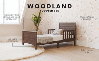 The Child Craft Woodland Toddler Bed in Brushed Truffle features pine construction, shiplap paneling, low-to-ground design, and built-in safety rails—ideal for a stylish nursery with floral wallpaper and plenty of natural light.