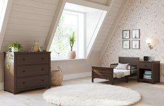 A cozy attic bedroom with sloped ceilings, a window, dark wood furniture including the Child Craft Harmony 3-Drawer Dresser with Changing Table Topper in Brushed Truffle, a toddler bed, nightstand, white round rug, plant, and decorative wall art and wallpaper.