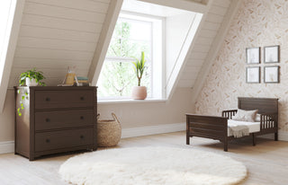 A cozy, light-filled attic nursery features a Child Craft Woodland Toddler Bed in Brushed Truffle, a dark wood dresser, plants, a woven basket, framed art, and a round white rug on a light wood floor.