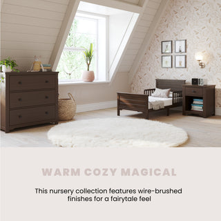 A cozy nursery features the Child Craft Woodland Toddler Bed in Brushed Truffle, wooden furniture, and soft lighting. A round rug and slanted windows create a warm, inviting space with a fairytale feel.