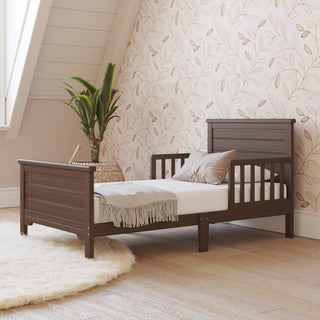 The Child Craft Woodland Toddler Bed in Brushed Truffle with a white mattress, grey blanket, and brown pillow sits on a light wooden floor by a round cream rug. A potted plant and leaf-patterned wallpaper add charm to the cozy room.