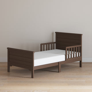 A Child Craft Woodland Toddler Bed in Brushed Truffle with side rails and a white mattress sits on a light wooden floor against a plain beige wall.
