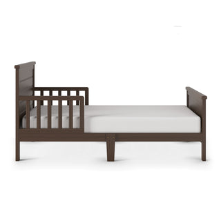 The Child Craft Woodland Toddler Bed in Brushed Truffle features a low-profile wooden frame, dark brown finish, slatted guardrails on one side, and includes a white mattress for a simple, sturdy sleep solution.