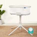 The HALO BassiNest® Soothing Swivel Sleeper 3.0 Bassinet, featuring mesh sides and a four-legged stand, sits on a woven rug. A green plant in a white pot is in the background with a Baby Safety Alliance Verified badge at the lower right.