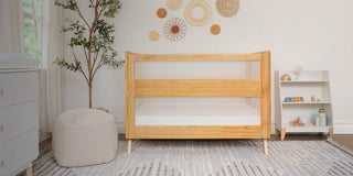 A cozy nursery featuring a wooden crib with white mesh panels, a pouf, and a small shelf with toys. Decorative wall hangings and a potted tree add warmth, while a soft rug covers the floor near a window with sheer curtains.