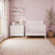 A pink-themed nursery showcases the Child Craft Atwood 4-in-1 Convertible Crib in Matte White, complemented by a matching dresser, a potted plant, and wall shelves filled with toys and books. Sunlight pours through a window adorned with curtains, brightening the room. A patterned rug lies atop the wooden floor, and baby-safe finishes throughout create a gentle environment for your little one.