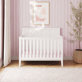 A Child Craft Atwood 4-in-1 Convertible Crib in Matte White, featuring baby-safe finishes and a pink mattress, stands against a pink striped wall. Abstract art hangs above the crib, while a potted plant rests on the wooden floor with a light rug nearby. Sunlight streams through a window with white curtains, highlighting the crib’s ability to later transform into a toddler bed.