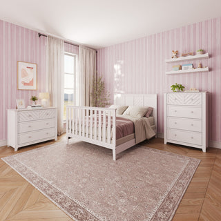 A bright bedroom with pink striped wallpaper features the Child Craft Atwood 4-in-1 Convertible Crib in Matte White, along with matching white dressers that have baby-safe finishes. Wall shelves provide extra storage, sheer curtains frame the sunlit window, and a large patterned rug covers the wooden floor.