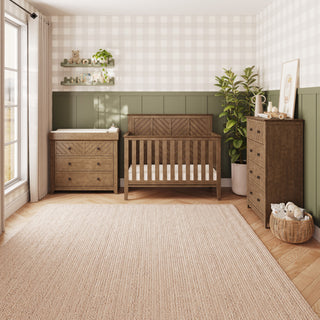 A cozy nursery features the Child Craft Atwood 4-in-1 Convertible Crib in Cocoa Bean, which can later transform into a toddler bed. The room also includes a dresser, changing table, and shelves filled with toys. A potted plant, framed picture, and a basket with blankets rest on the beige rug as sunlight streams in through the large window.