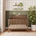 The Child Craft Atwood 4-in-1 Convertible Crib in Cocoa Bean is placed in a nursery with green and white walls, complemented by a potted plant. Above the crib, shelves display books, a teddy bear, and decorative items, while sunlight streams through curtains on the left.