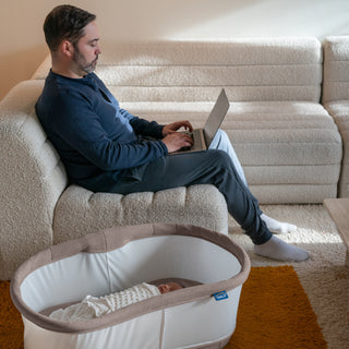 A man works on his laptop from a sofa while a baby sleeps nearby in the HALO BassiNest® Connected Swivel Sleeper 3.0 smart bassinet, which features 360° swivel, set on a rug in a cozy living room.