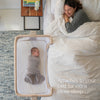 A smiling woman lies in bed under a white comforter, next to a HALO 2-in-1 Bedside Bassinet Sleeper with a swaddled baby inside. Text reads: Attaches to your bed for extra close sleep.