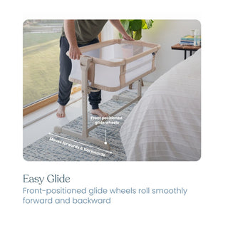 A person uses the HALO 2-in-1 Bedside Bassinet Sleeper in light brown and white with breathable mesh. Its front-positioned glide wheels let the bassinet smoothly slide forward and backward beside a bed, as shown by arrows.