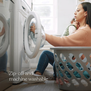 A woman sits on the floor with a child, loading laundry into a front-loading washer. A HALO 2-in-1 Bedside Bassinet Sleeper is nearby. Text reads: Zip-off and machine washable.
