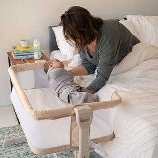 A woman tends to her baby in a HALO Bedside Sleeper Bassinet Bundle. A nearby nightstand holds books, a bottle, and baby essentials, creating a calm and cozy scene.
