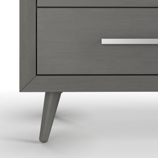 Close-up of the bottom corner of the Cranbrook Double Dresser in Lunar Gray by Child Craft, featuring angled legs and a sleek silver drawer handle—a stylish addition to any nursery furniture set.