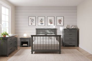 A modern bedroom features gray wooden furniture, including a bed, dresser, nightstand, and the Denman 4 Drawer Chest by Child Craft in Midnight Gray. Decor includes botanical art, a plant, a lamp, neutral walls, and light wood floors.