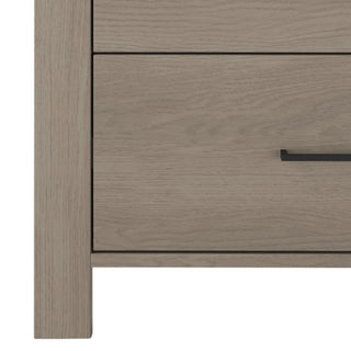 Close-up of the Child Craft Denman 4 Drawer Chest in Crescent Gray with a modern black handle, two visible drawers, and part of the left leg. The wood grain highlights the natural, stylish look of this nursery furniture piece.
