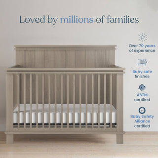 The Denman 2-Piece Crib and DreamWeave Set in Crescent Grey by Child Craft sits in a bright room. Above, text reads Loved by millions of families. At right, icons show 70+ years' experience, baby-safe finishes, ASTM & Baby Safety Alliance certified.