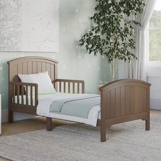 The Child Craft Hampton 2-Piece Toddler Bed and DreamWeave Set in Dusty Heather features a high headboard and footboard, set on a light rug in a room with star-patterned wallpaper, sheer curtains, white bedding, green blanket, and a potted tree.