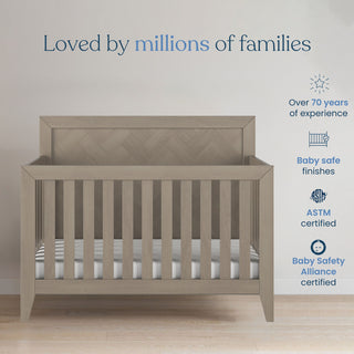 A Kieran 3 Piece Nursery Set with Crib, Dresser, and Chest by Child Craft is showcased in a room, with the gray wooden convertible crib featuring a striped mattress that beautifully complements any nursery furniture set. Above, the text reads, "Loved by millions of families." On the right, blue icons and text highlight safety certifications and years of experience.