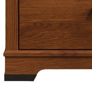 Close-up of the bottom corner of the Child Craft Redmond 4 Drawer Chest in Coach Cherry, featuring visible wood grain and a rich cherry finish. The base has a simple, gently curved design with a dark accent at the foot.