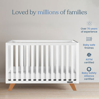 A sleek matte white Child Craft Soho 2-Piece Crib and DreamWeave Set with wooden legs stands on a light wood floor. Text above: Loved by millions of families. Icons highlight safety, 70+ years’ experience, and DreamWeave mattress compatibility.