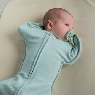 A baby in a light blue HALO SwaddleStretch Newborn Swaddle Bundle lies on a soft cream-colored surface, turned slightly to the side and sucking on a green pacifier.