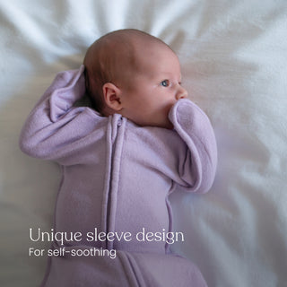 A baby lies on a white blanket, wearing a light purple HALO SwaddleStretch™ Newborn Swaddle TOG 0.5 with a unique sleeve design for self-soothing. Text on the image: Unique sleeve design. For self-soothing.