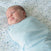 A baby in a light blue HALO Swaddle 100% Organic Cotton TOG 1.5 rests on a pastel floral sheet, looking to the side—perfect for healthy hip development.