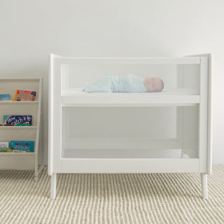 A baby sleeps soundly in a white crib with mesh sides, wrapped in the HALO Swaddle 100% Organic Cotton TOG 1.5 for healthy hip development. A bookshelf of children’s books and a textured rug complete the serene, neutral room.