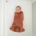 A newborn is snugly wrapped in a HALO Swaddle Plushy Dot Velboa TOG 3.0, textured and rust-colored, lying peacefully on a white crib mattress.