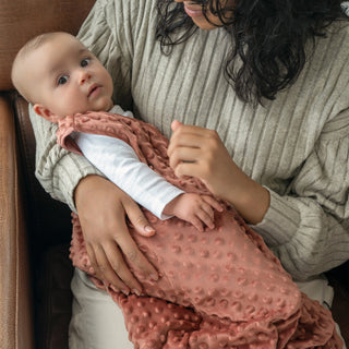 A baby rests calmly in a woman’s arms, wrapped in the HALO Wearable Blanket Plushy Dot Velboa TOG 1.5 in soft pink. The woman wears a light gray ribbed sweater and sits on a brown couch, showcasing safe and cozy sleep.
