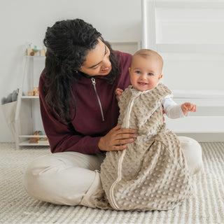 A smiling woman sits cross-legged, holding a happy baby dressed in the HALO Wearable Blanket Plushy Dot Velboa TOG 1.5. They’re in a bright, cozy room with a soft rug and shelves, creating an ideal space for safe sleep.
