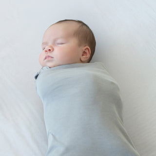 A newborn baby sleeps peacefully, swaddled in a light grey HALO Swaddle 100% Organic Cotton TOG 1.5 on a white surface.