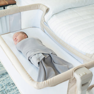 A baby sleeps peacefully in a beige bassinet beside a bed with a white quilt, wrapped in a HALO Swaddle 100% Organic Cotton TOG 1.5, lying on their back for a safe and cozy rest.