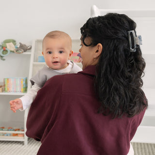A woman with dark hair in a clip holds a baby wearing the HALO Wearable Blanket 100% Organic Cotton TOG 0.5, both looking at the camera in a softly lit room filled with children’s toys and books, creating a cozy, safe sleep space.