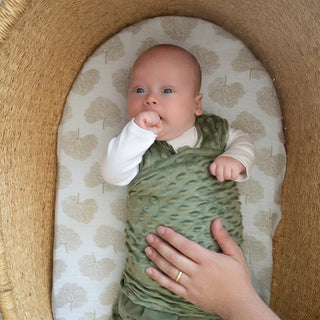 A baby lies in a woven bassinet, snug in a green HALO Swaddle Plushy Dot Velboa TOG 3.0, with one hand near their mouth. An adult hand with a gold ring gently rests on the baby's plush adjustable swaddle blanket.
