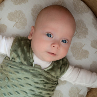 A baby with blue eyes and light skin, wearing a green vest over a white shirt, lies on a beige leaf-patterned cushion looking up with arms outstretched in the HALO Swaddle Plushy Dot Velboa TOG 3.0 for comfort.